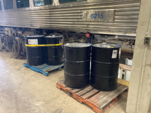 Colour photograph showing black barrels on pallets next to a stainless steel rail car.