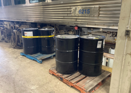 Colour photograph showing black barrels on pallets next to a stainless steel rail car.